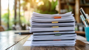 A stack of papers on a desk with sunlight coming through a window in the background.